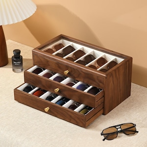 May include: A dark wood sunglasses storage organizer with a glass top lid and two pull-out drawers. The interior features white velvet-lined compartments for multiple pairs of eyewear. Each drawer has a small gold-toned knob handle.