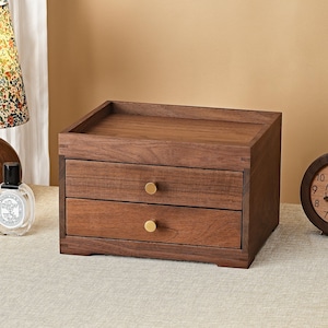 May include: A rectangular wooden storage organizer featuring two horizontal drawers with small, round, gold-toned metal knobs. The unit has a flat top surface with raised edges, showcasing a natural dark brown wood grain finish throughout the structure.