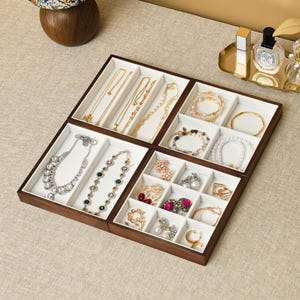 May include: A dark wood jewelry organizer tray with a white velvet interior, divided into multiple compartments. It contains various necklaces, bracelets, earrings, and rings, including crystal-embellished pieces, gold chains, and pearl strands.