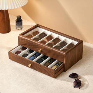 May include: A rectangular wooden storage box with a glass lid and a pull-out drawer, featuring multiple white-lined compartments for organizing sunglasses. The box has two small gold-toned knobs and contains several pairs of sunglasses.