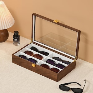 May include: A rectangular wooden storage box with a clear glass lid and a gold-tone metal latch. The interior features white dividers creating six individual compartments, each containing a pair of sunglasses with dark frames and lenses.