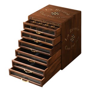 May include: A dark brown wooden jewelry chest with six pull-out drawers featuring glass fronts and gold-toned metal handles. The top and side are engraved with the text La Reverie Cree Par M&m and a small checkered square emblem. The interior is lined.