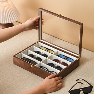 May include: A rectangular wooden storage box with a hinged glass lid and a gold-tone metal latch. The interior is divided into eight individual compartments lined with off-white fabric, each containing a pair of sunglasses with various frame colors.
