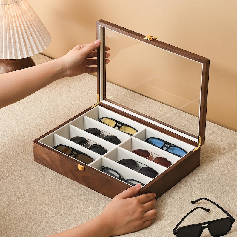 May include: A rectangular wooden storage box with a hinged glass lid and a gold-tone metal latch. The interior is divided into eight individual compartments lined with off-white fabric, each containing a pair of sunglasses with various frame colors.