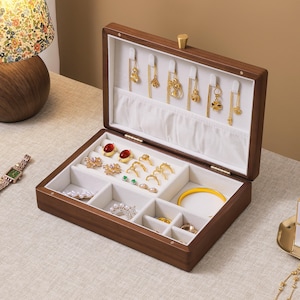 May include: A rectangular wooden jewelry box with a dark brown finish and a white interior lining. The lid features hooks for hanging necklaces, while the base contains multiple compartments for rings, earrings, and bracelets, filled with gold jewelry.