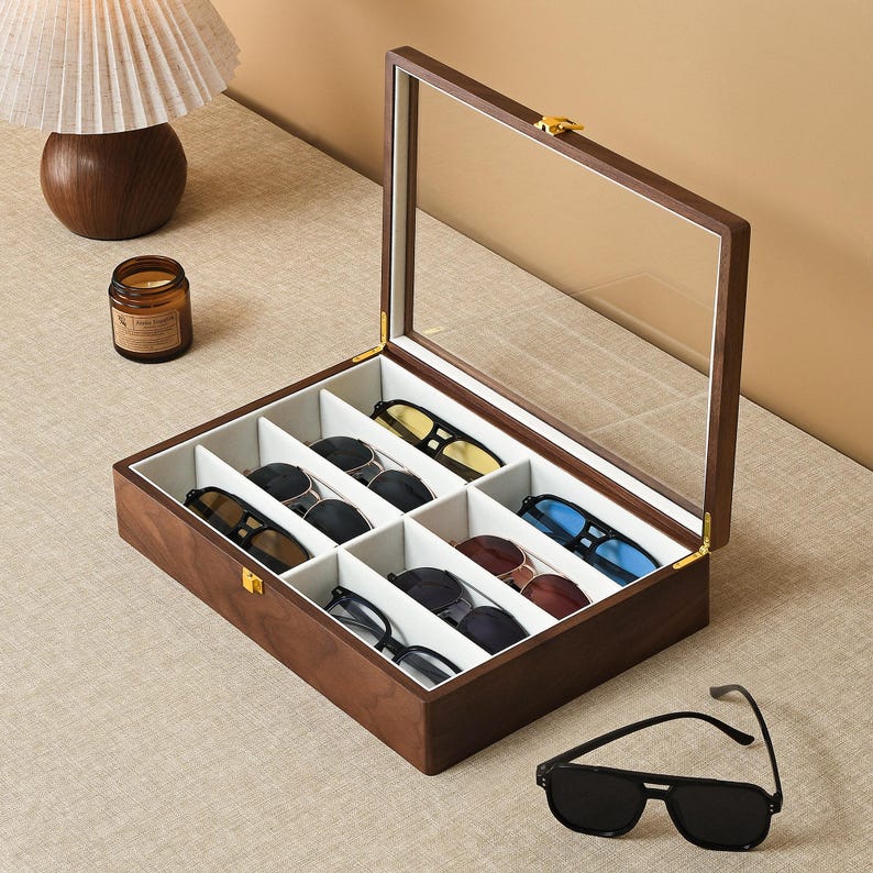 May include: A rectangular wooden storage box with a hinged glass lid and a gold-toned metal latch. The interior is divided into six white-lined compartments, each containing a pair of sunglasses. A separate pair of black sunglasses rests nearby.