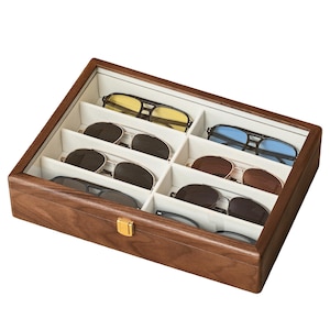 May include: A rectangular wooden storage box with a glass lid and a gold-colored metal latch, featuring eight interior compartments lined with cream-colored fabric, each containing a pair of sunglasses with various lens colors and frame styles.