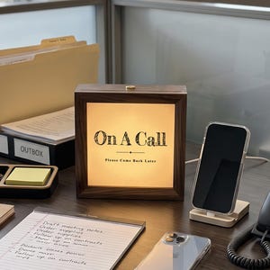 May include: A square wooden light box with a dark brown frame and a glowing cream-colored center panel. The text "On A Call" is printed in black, followed by a horizontal line and the smaller text "Please Come Back Later".