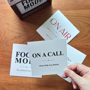 May include: A set of square white cards with black and red text. One card reads ON A CALL I'll be With You Shortly, another reads ON AIR SHIFT YOUR S, and a third reads FOCUS MODE WorkflowCon. A wooden stand displays a FOCUS MODE card.