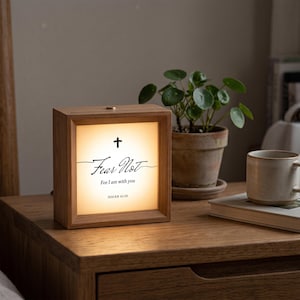 May include: A square wooden light box with a warm glow, featuring a black cross icon and the text "Fear Not For I am with you ISAIAH 41:10" printed on the front panel. The frame is made of light-toned wood with a smooth finish and a small button on top.