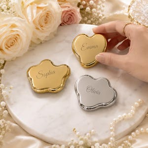 May include: Three small, irregular-shaped metal compact mirrors with polished finishes. Two are gold-toned, engraved with the names Emma and Sophia, and one is silver-toned, engraved with the name Olivia. Each piece features a smooth, reflective surface.