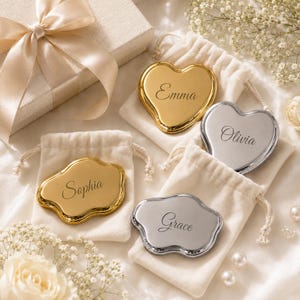 May include: Four metallic pocket mirrors in heart and cloud shapes, finished in gold and silver tones. Each mirror is engraved with a name: Emma, Olivia, Sophia, or Grace. The mirrors are placed on individual cream-colored fabric drawstring pouches.