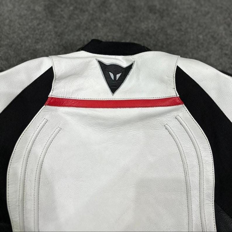 K&ouml;nnte beinhalten: Wei&szlig;-schwarze Motorradjacke mit rotem Streifen auf dem R&uuml;cken. Die Jacke hat einen schwarzen Kragen und ein Logo auf dem oberen R&uuml;cken. Die Jacke hat ein gestepptes Design auf dem R&uuml;cken.