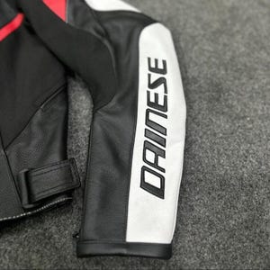 K&ouml;nnte beinhalten: Nahaufnahme eines schwarzen und wei&szlig;en Motorradjacken&auml;rmels aus Leder mit dem Markennamen "DAINESE" in gro&szlig;en, fetten Buchstaben aufgedruckt. Die Jacke hat rote Akzente und einen schwarzen Lederriemen.