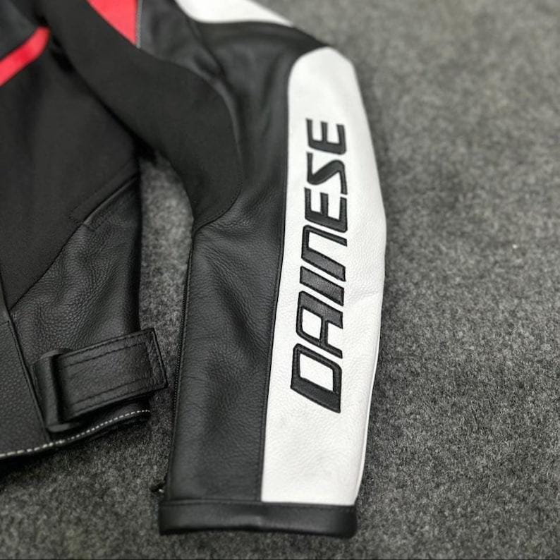 K&ouml;nnte beinhalten: Nahaufnahme eines schwarzen und wei&szlig;en Motorradjacken&auml;rmels aus Leder mit dem Markennamen "DAINESE" in gro&szlig;en, fetten Buchstaben aufgedruckt. Die Jacke hat rote Akzente und einen schwarzen Lederriemen.