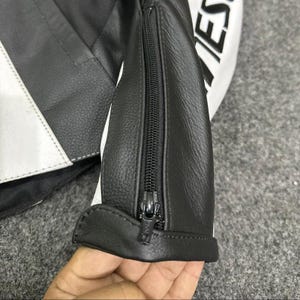 K&ouml;nnte beinhalten: Nahaufnahme eines schwarzen und wei&szlig;en Motorradjacken&auml;rmels aus Leder mit Rei&szlig;verschluss. Die Jacke hat wei&szlig;e Streifen und das Wort "IES" ist auf dem &Auml;rmel zu sehen.