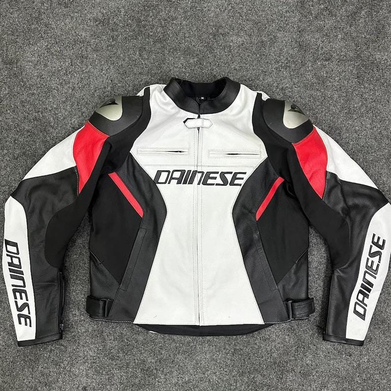 K&ouml;nnte beinhalten: Schwarze, wei&szlig;e und rote Motorradjacke aus Leder mit dem Markennamen "DAINESE" auf Brust und &Auml;rmeln. Die Jacke hat einen Stehkragen, Schulterpolster und einen Tailleng&uuml;rtel. Die Jacke ist auf Schutz und Stil ausgelegt.