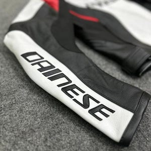 K&ouml;nnte beinhalten: Nahaufnahme eines schwarzen und wei&szlig;en Motorradjacken&auml;rmels aus Leder. Der &Auml;rmel zeigt das Wort "DAINESE" in fetten, schwarzen Buchstaben auf einem wei&szlig;en Streifen. Rote Akzente sind im Schulterbereich der Jacke zu sehen. Die Jacke scheint aus hochwertigem Leder zu sein.