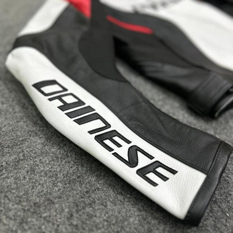 K&ouml;nnte beinhalten: Nahaufnahme eines schwarzen und wei&szlig;en Motorradjacken&auml;rmels aus Leder. Der &Auml;rmel zeigt das Wort "DAINESE" in fetten, schwarzen Buchstaben auf einem wei&szlig;en Streifen. Rote Akzente sind im Schulterbereich der Jacke zu sehen. Die Jacke scheint aus hochwertigem Leder zu sein.