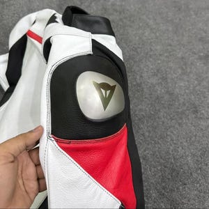 K&ouml;nnte beinhalten: Nahaufnahme einer Motorradjacke aus wei&szlig;em, schwarzem und rotem Leder. Die Jacke hat einen schwarzen Schulterbereich aus Leder mit einem silbernen Logo. Die Jacke hat einen roten Einsatz am Unterarm und wei&szlig;e Eins&auml;tze am Oberarm und am Oberk&ouml;rper.