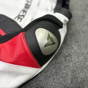 K&ouml;nnte beinhalten: Nahaufnahme eines Details einer Motorradjacke aus wei&szlig;em, schwarzem und rotem Leder. Die Jacke hat ein silbernes Logo am Ellbogen, wobei der Markenname teilweise sichtbar ist. Das Design umfasst rote und schwarze Akzente.