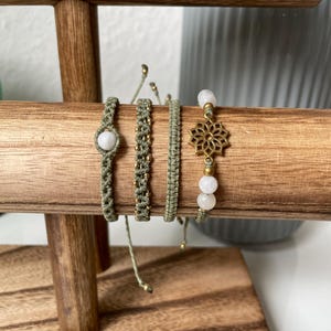 Puede incluir: Cuatro pulseras de macramé hechas a mano en tonos verdes y dorados se exhiben en un soporte de madera. Las pulseras presentan varios diseños, incluyendo una piedra blanca central, detalles de cuentas y un dije de filigrana dorada con cuentas blancas.