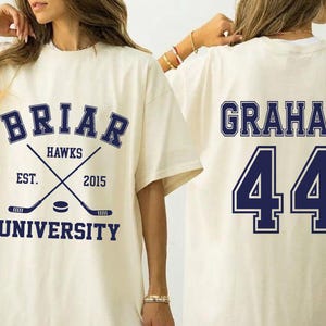 以下が含まれることがあります： クリーム色のTシャツ。前面には「BRIAR HAWKS UNIVERSITY」の文字と交差したホッケーのスティックのネイビーブルーのグラフィック。背面には「GRAHAM」と数字の「44」がネイビーブルーで表示されています。