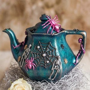 Puede incluir: Una tetera color verde azulado decorada con telarañas plateadas, perlas blancas, arañas rosas y figuras de esqueletos plateados. La tapa tiene una araña rosa. La tetera está rodeada por una rosa blanca, una llave pequeña y un libro en miniatura.