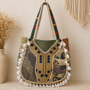 Può includere: Una borsa tote colorata con un design patchwork. La borsa presenta una varietà di tessuti fantasia nei toni del verde, dell'oro e del grigio, con nappe bianche lungo i bordi. I manici sono color crema con un motivo floreale.