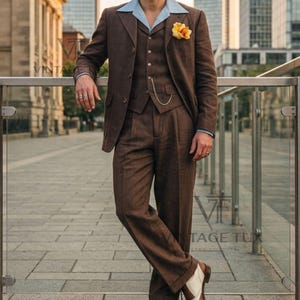 May include: A brown three-piece suit consisting of a blazer, a vest with a silver chain, and trousers, paired with a light blue collared shirt, a grey flat cap, and two-tone brown and white leather dress shoes with a yellow flower lapel pin.