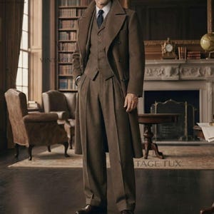 May include: A brown wool three-piece suit consisting of a long overcoat, a buttoned vest, and tailored trousers. The ensemble includes a white collared shirt and a dark tie. The individual wears dark sunglasses and brown leather dress shoes.