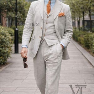 May include: A light beige three-piece linen suit consisting of a blazer, a vest, and trousers. The outfit includes a light blue button-down shirt, an orange necktie, a matching pocket square, a fedora hat with a brown band, and two-tone shoes.