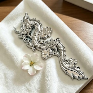 May include: A detailed silver dragon figurine with cloud motifs, set against a white backdrop. The dragon's scales and features are clearly defined. A white flower with pink tips is also present. The figurine has a shiny finish.
