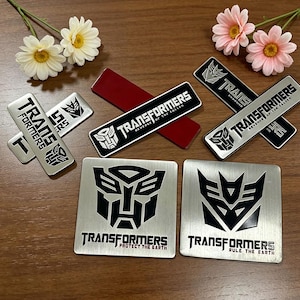 Könnte beinhalten: Eine Sammlung silberner und schwarzer Transformers-Emblem-Aufkleber und -Abzeichen. Die Aufkleber zeigen das Transformers-Logo und den Text, wobei einige den Satz "Revenge of the Fallen" und "Protect the Earth" enthalten.