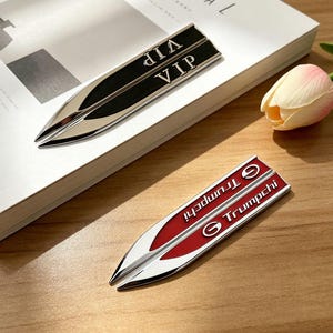 May include: Two metallic car emblems. One is black with "VIP" in silver lettering. The other is red with "Trumpchi" in white lettering. Both have a pointed, arrow-like design.