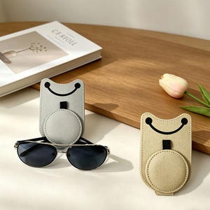 Peut inclure: Deux étuis à lunettes de soleil gris clair et beige, chacun avec un motif de visage souriant. Les étuis ont une base circulaire et une boucle pour attacher à un téléphone. Une paire de lunettes de soleil repose devant l'étui gris.