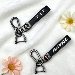 Könnte beinhalten: Zwei schwarze Leder-Schlüsselanhänger mit Metallbeschlägen. Einer trägt die Aufschrift "VIP" in weißen Buchstaben, der andere "HAVAL". Beide haben einen Metallkarabiner und einen D-Ring für Schlüssel.