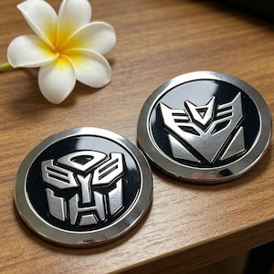 Puede incluir: Dos emblemas redondos, plateados y negros, con los logotipos de Transformers. Uno presenta el símbolo Autobot, el otro el símbolo Decepticon. Una flor blanca y amarilla en el fondo.