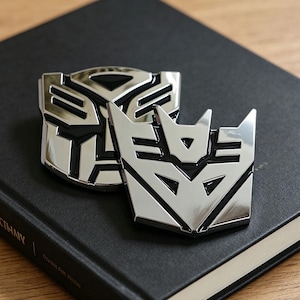 May include: Two silver Transformers logo emblems with black detailing. The emblems are set against a dark background, highlighting their shiny, metallic appearance. The logos are a well-known symbol from the Transformers franchise.