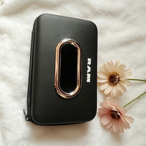 May include: Black rectangular case with a gold-colored oval accent and the word "RAM" in white. The case has a zipper closure and is next to two daisies with pink and white petals.