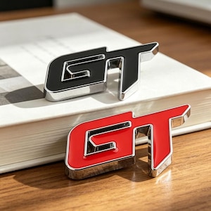May include: Two metallic GT emblems, one black and one red, with a silver border. The emblems are displayed on a white book and a wooden surface. The GT emblems are likely for automotive use.