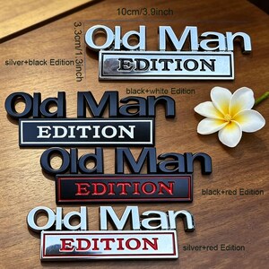 Może przedstawiać: Kolekcja metalowych emblemat&oacute;w samochodowych z napisem "Old Man EDITION" w r&oacute;żnych kombinacjach kolorystycznych. Emblematy mają około 10 cm długości i 3,3 cm wysokości. Opcje kolorystyczne obejmują srebrny z czarnym, czarny z białym, czarny z czerwonym i srebrny z czerwonym.