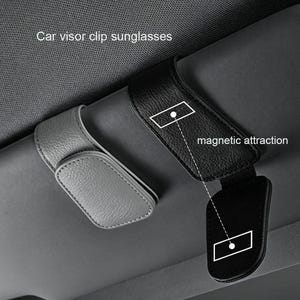 Könnte beinhalten: Ein Sonnenbrillen-Clip für die Autovisierung, in Grau und Schwarz. Der Clip verfügt über eine magnetische Anziehungskraft, wie durch den Text angezeigt. Das Produkt dient dazu, Sonnenbrillen sicher im Fahrzeug zu halten.
