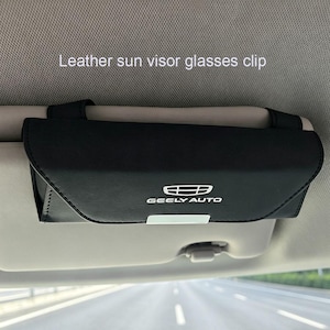 May include: A black leather glasses clip for a car sun visor, with the text "Leather sun visor glasses clip" and the "GEELY AUTO" logo. The clip is attached to the sun visor.