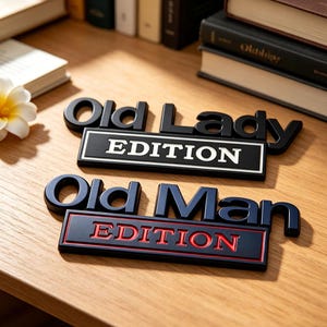 Może przedstawiać: Dwa czarno-niebieskie znaki dekoracyjne z napisami "Old Lady EDITION" w kolorze białym i "Old Man EDITION" w kolorze czerwonym. Znajdują się na drewnianej powierzchni, z książkami w tle.