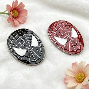 Puede incluir: Dos broches ovalados con la cara de Spider-Man. Uno es negro con un diseño de telaraña blanca y formas de ojos blancos. El otro es rojo con un diseño de telaraña blanca y formas de ojos blancos. Los broches están sobre una superficie blanca y esponjosa.