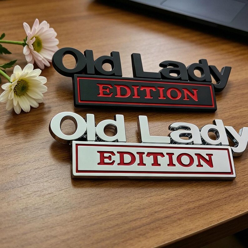 Może przedstawiać: Dwa metalowe emblematy samochodowe z napisem "Old Lady EDITION". Jeden jest czarny z czerwonymi literami, drugi srebrny z czerwonymi literami. Leżą na drewnianej powierzchni, z kwiatami w tle.