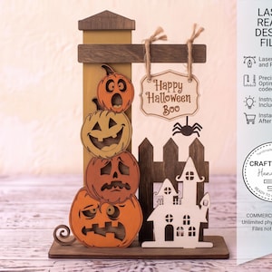 Może przedstawiać: Wycinana laserowo drewniana dekoracja na Halloween przedstawiająca stos czterech pomarańczowych dyń o różnych wyrazach twarzy, ciemnobrązowy płot, białą sylwetkę nawiedzonego domu oraz wiszący znak z napisem Happy Halloween Boo.