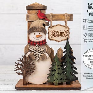 May include: A wooden snowman ornament holding a snowflake, wearing a hat and scarf, and standing in front of a fence and trees. A sign that says "BELIEVE" hangs above the snowman. A red bird sits on top of the sign. The words "LASER READY DESIGN FILES" are on the right.