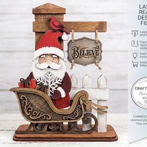 May include: A wooden Christmas decoration featuring Santa Claus in a sleigh, holding a snowflake and gifts. A sign that says "BELIEVE" hangs above a white picket fence. A red bird sits atop a wooden sign. The words "LASER READY DESIGN FILES" are visible.